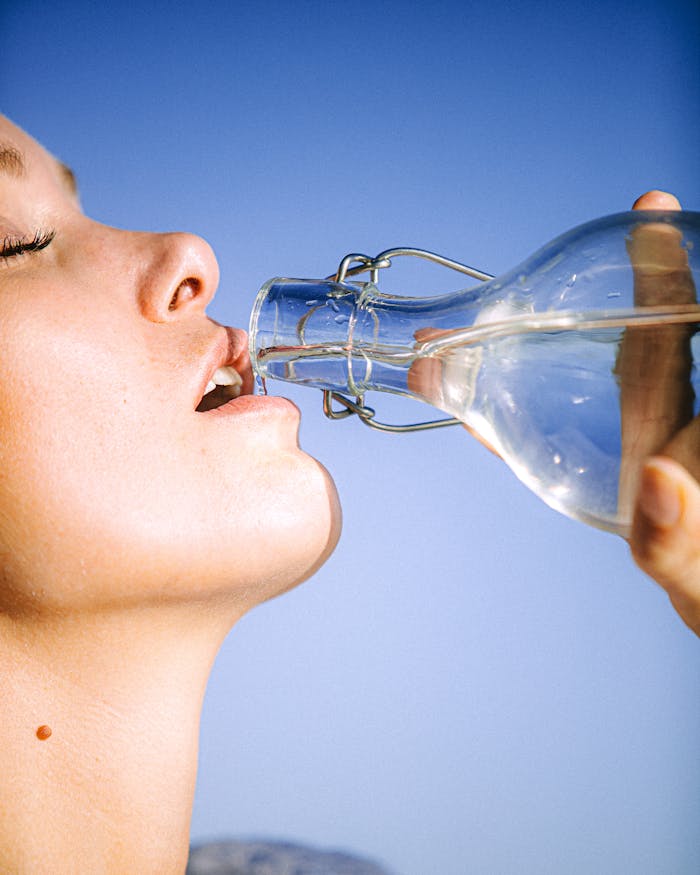 A woman drinking water from a glass bottle against a clear blue sky, emphasizing hydration.